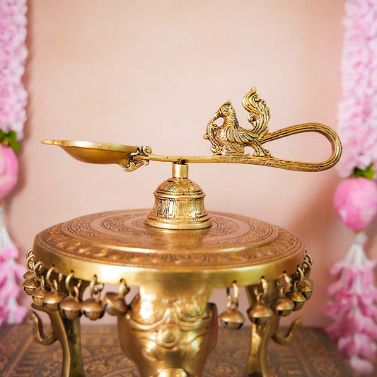 Peacock-shaped brass Aarathi lamp with a detailed handle and ornate carvings, displayed on a decorative surface.