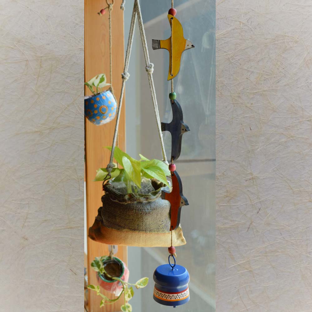 Hand-painted wooden flying birds in yellow, blue, and orange with a colorful blue metal bell wind chime, hanging with rustic jute rope and wooden beads near a window with greenery.