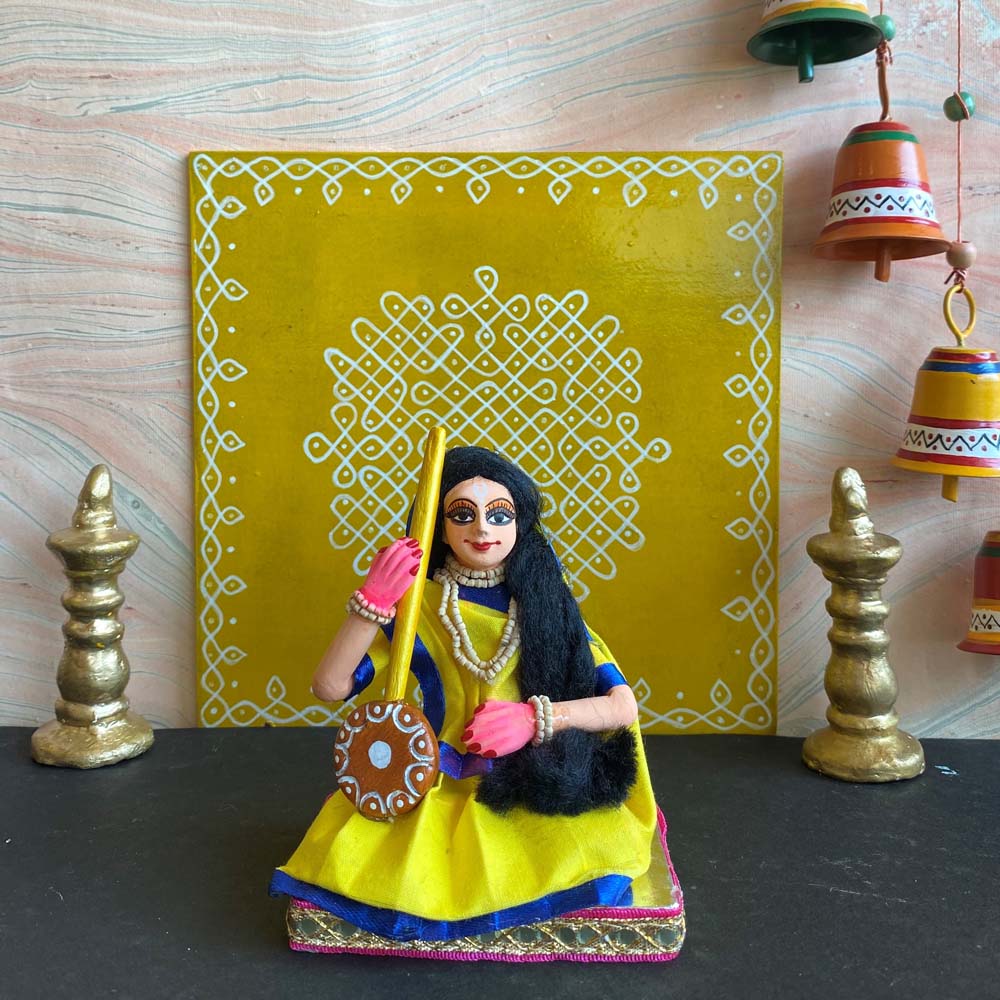 Handcrafted Navaratri costume doll of Meerabai dressed in yellow and blue, holding a tambura, placed against a decorative yellow kolam backdrop.