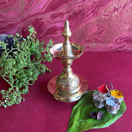 Handcrafted brass Udupi oil lamp with intricate carvings, placed on a red surface with pooja items and greenery.