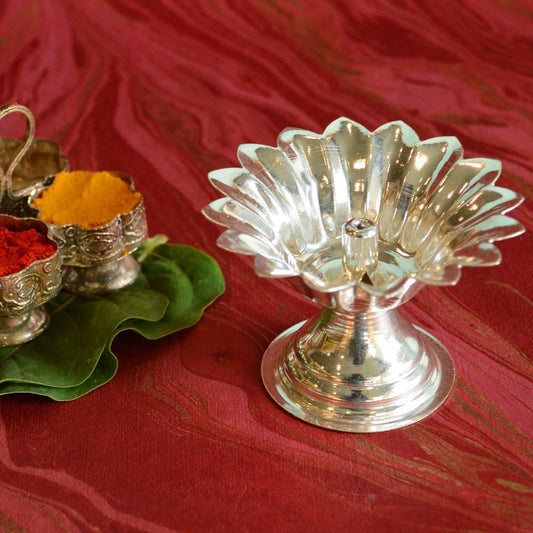 Handcrafted silver-finish Chandan Flower oil lamp with intricate floral design, placed on a red surface with pooja items and green leaves.