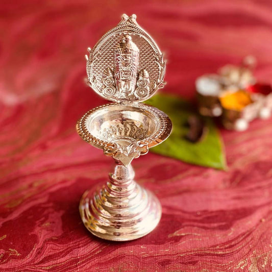 Handcrafted silver-finish Balaji oil lamp with intricate detailing, placed on a red surface with greenery and pooja items in the background.