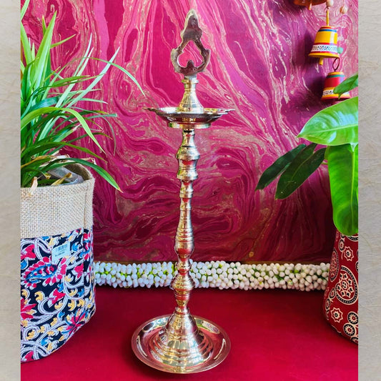 Traditional brass kuthu vilakku (oil lamp) with an intricate top design, surrounded by plants and vibrant decor on a red surface.