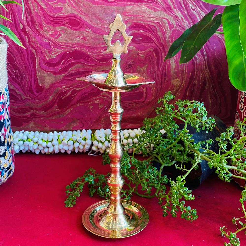 Traditional brass kuthu vilakku (oil lamp) with an intricate top design, surrounded by plants and vibrant decor on a red surface.