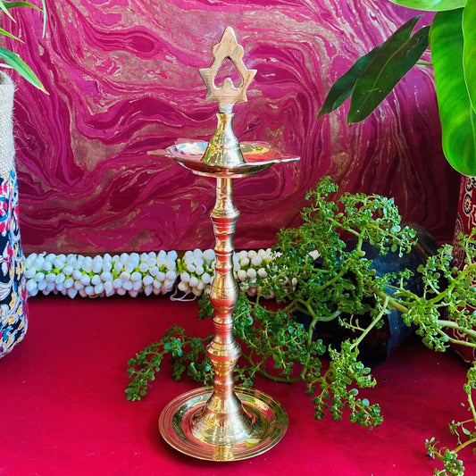 Traditional brass kuthu vilakku (oil lamp) with an intricate top design, surrounded by plants and vibrant decor on a red surface.