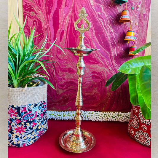 Traditional brass kuthu vilakku (oil lamp) with an intricate top design, surrounded by plants and vibrant decor on a red surface.