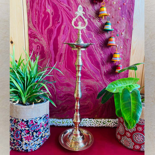 Traditional brass kuthu vilakku (oil lamp) with an intricate top design, surrounded by plants and vibrant decor on a red surface.