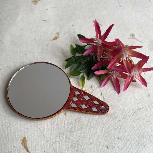 Handheld round decorative mirror with a red glitter handle and geometric accents, ideal for pooja, home decor, or grooming.