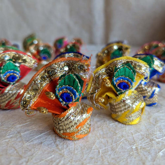 Colorful handcrafted decorative turbans with peacock feather motifs, zari accents, and sequins, displayed on a textured background.