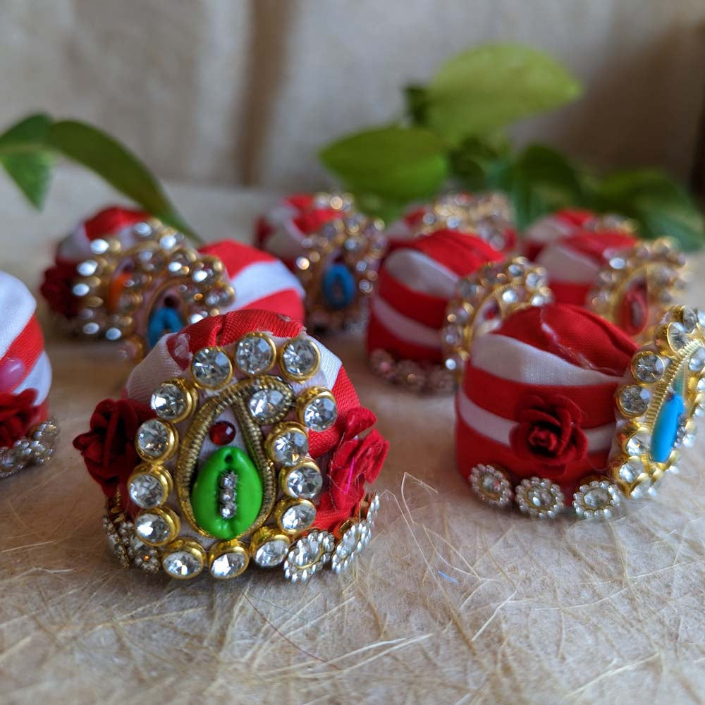 Handcrafted red and white decorative turbans adorned with crystals, floral accents, and colorful centerpiece details, displayed on a textured surface.