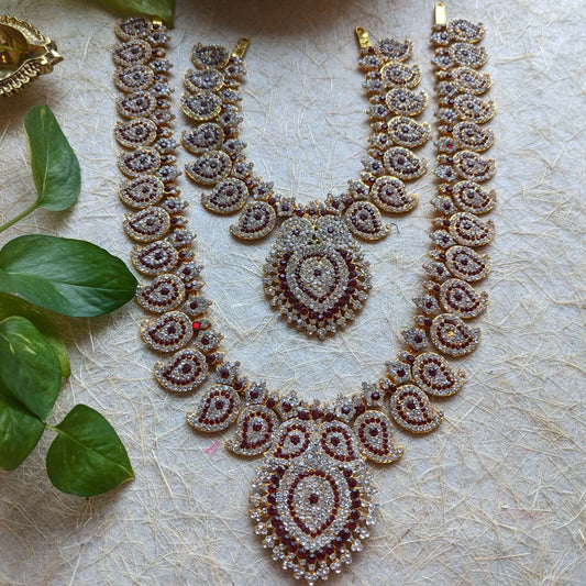 Gold-plated long necklace set with paisley design, multi-colored stones, and a central pendant, placed on a textured surface with green leaves.