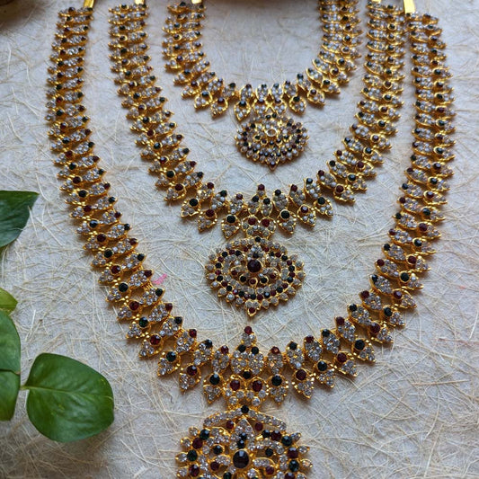 Gold-plated long necklace set with multi-colored stones, featuring intricate designs, placed on a textured surface with green leaves.