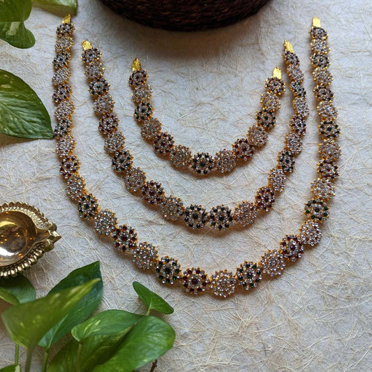 Gold-plated long necklace set with floral motifs and multi-colored stones, placed on a textured surface with green leaves and a brass diya.