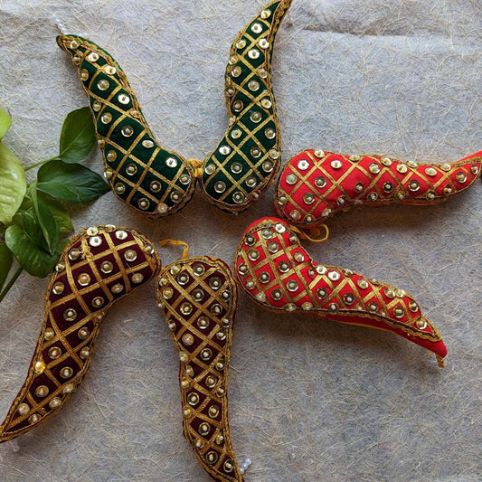 Handcrafted Vaagamalai garland in red, green, and maroon with gold latticework and mirror embellishments, placed on a textured surface with green leaves.