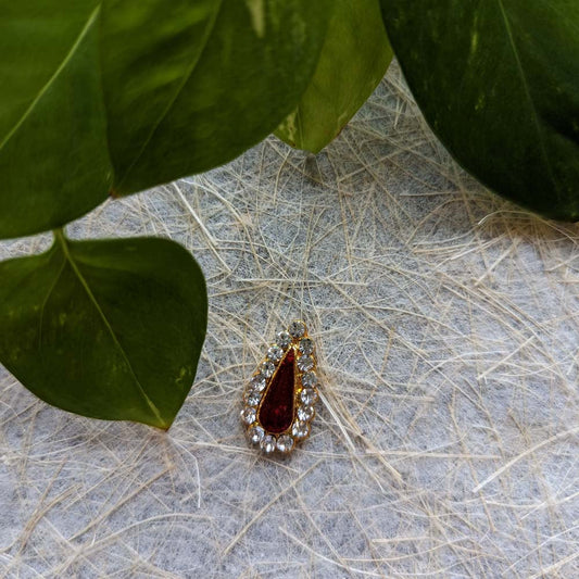 Gold-plated Small Tilak Ornament with a red teardrop centerpiece and white stones, placed on a textured surface with green leaves.