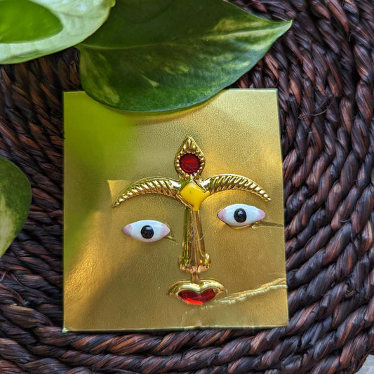 Traditional deity face decoration with ruby accents and gold-toned details, displayed on a woven mat with green leaves.