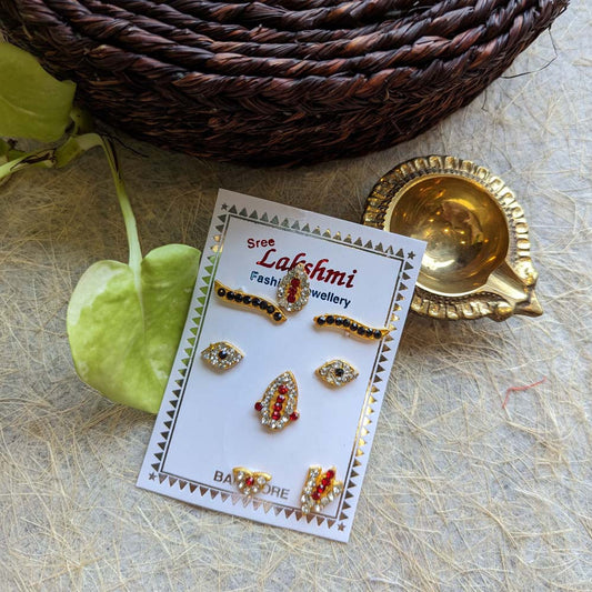 Traditional deity face decoration set with crystal and ruby embellishments, displayed on a decorative card with brass diya and leaves.