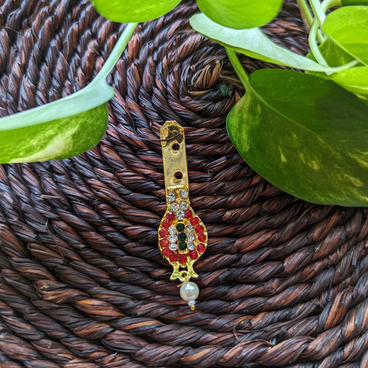 Traditional gold-toned Nethi Chutti with crystal, ruby, and emerald embellishments, and a pearl drop, displayed on a woven brown mat with green leaves.