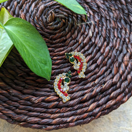 Crescent-shaped studded earrings with red, green, and white stones on a gold-toned base, placed on a woven background with green leaves.