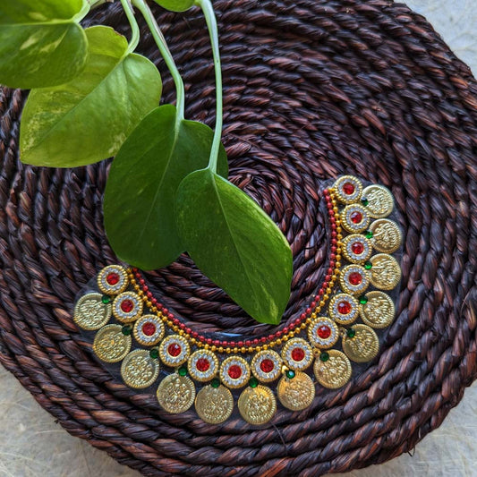 Traditional sticker necklace with gold coin motifs and red-green embellishments, displayed on a woven surface with green leaves.