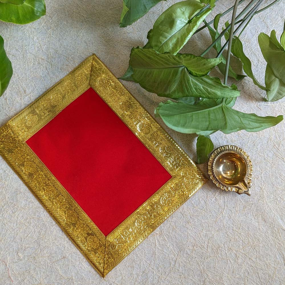 Red velvet aasan with a golden zari border, paired with a brass lamp, displayed on a textured surface with green leaves in the background.