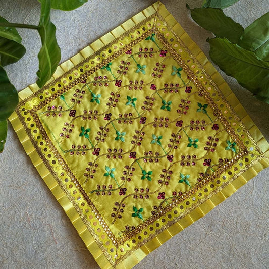 A gold 11x11 embroidered Aasan with intricate floral patterns, sequin embellishments, and a golden decorative border, displayed with green leaves.