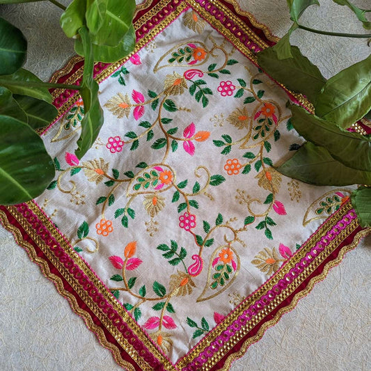 A 14x14 embroidered Aasan with floral patterns in vibrant colors, accented by a pink and gold decorative border, displayed alongside green leaves.